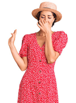 Young beautiful woman wearing summer hat smelling something stinky and disgusting, intolerable smell, holding breath with fingers on nose. bad smell