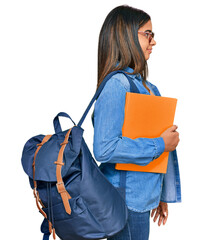 Young latin girl wearing student backpack and holding books looking to side, relax profile pose...