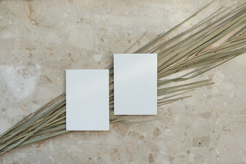 Blank paper invitation cards with copy space. Dried tropical palm leaf stem on marble stone background. Flat lay, top view aesthetic minimalist wedding invitations