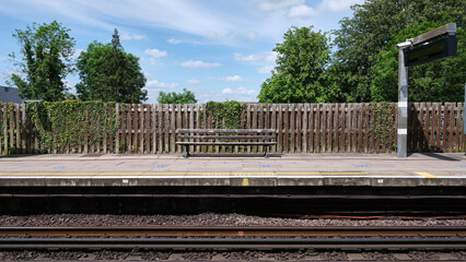 Train station platform with a wooden bench and fence. Trees and blue sky with white clouds in the background.