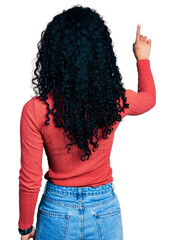 Young hispanic woman with curly hair wearing glasses posing backwards pointing ahead with finger hand