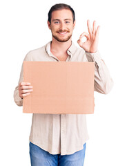 Young handsome caucasian man holding empty cardboard banner doing ok sign with fingers, smiling friendly gesturing excellent symbol