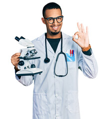 Young african american man wearing scientist uniform holding microscope doing ok sign with fingers, smiling friendly gesturing excellent symbol