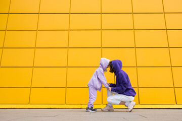 Woman in purple hoody have fun with cute girl 4-5 years old. Mommy and little daughter on yellow mall wall background. Childhood concept of love for family and fatherhood on Mother's Day