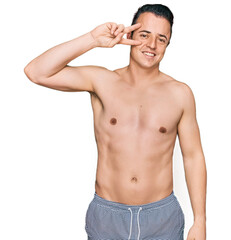 Fototapeta premium Handsome young man wearing swimwear shirtless doing peace symbol with fingers over face, smiling cheerful showing victory