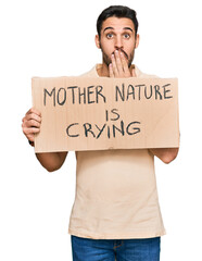 Young hispanic man holding mother nature is crying protest cardboard banner covering mouth with hand, shocked and afraid for mistake. surprised expression