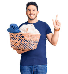Young hispanic man holding laundry basket surprised with an idea or question pointing finger with...