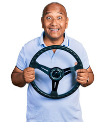 Hispanic middle age man holding steering wheel celebrating crazy and amazed for success with open...