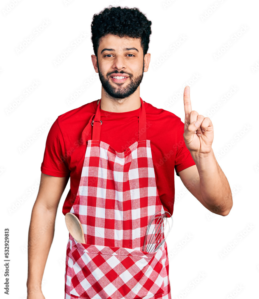 Sticker young arab man with beard wearing cook apron showing and pointing up with finger number one while sm