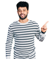 Young arab man with beard wearing casual striped sweater with a big smile on face, pointing with hand finger to the side looking at the camera.