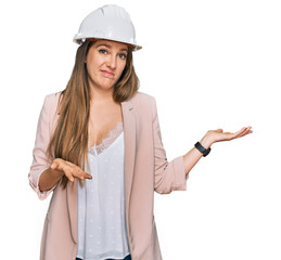 Young blonde woman wearing architect hardhat clueless and confused expression with arms and hands raised. doubt concept.