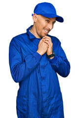 Bald man with beard wearing builder jumpsuit uniform laughing nervous and excited with hands on chin looking to the side