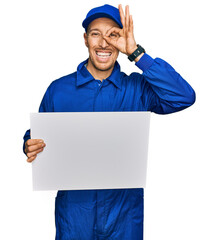 Bald man with beard wearing builder jumpsuit uniform holding empty banner smiling happy doing ok sign with hand on eye looking through fingers