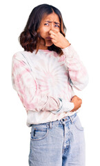 Young beautiful mixed race woman wearing casual tie dye sweatshirt smelling something stinky and...