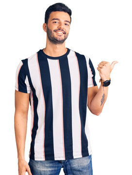 Young handsome hispanic man wearing striped tshirt smiling with happy face looking and pointing to the side with thumb up.