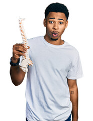 Young african american man holding anatomical model of spinal column scared and amazed with open...