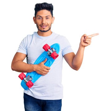 Handsome latin american young man holding skate smiling happy pointing with hand and finger to the side
