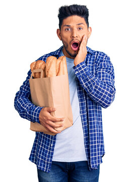 Handsome latin american young man holding paper bag with bread afraid and shocked, surprise and amazed expression with hands on face
