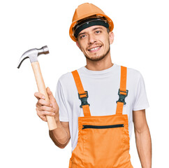 Hispanic young man wearing hardhat holding hammer looking positive and happy standing and smiling with a confident smile showing teeth