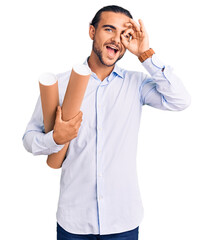 Young handsome man holding paper blueprints smiling happy doing ok sign with hand on eye looking through fingers