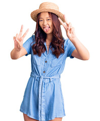 Obraz na płótnie Canvas Young beautiful chinese girl wearing summer hat smiling with tongue out showing fingers of both hands doing victory sign. number two.
