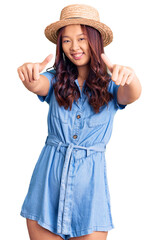 Fototapeta premium Young beautiful chinese girl wearing summer hat approving doing positive gesture with hand, thumbs up smiling and happy for success. winner gesture.