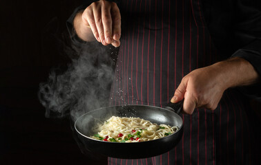 The cook adds salt to the spaghetti in a steaming hot pan. Recipe or menu for restaurant or hotel on black background.