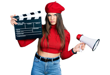 Young brunette teenager holding video film clapboard and megaphone skeptic and nervous, frowning...