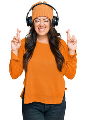 Fototapeta premium Beautiful brunette young woman listening to music using headphones gesturing finger crossed smiling with hope and eyes closed. luck and superstitious concept.