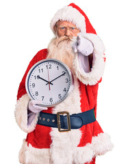 Old senior man with grey hair and long beard wearing santa claus costume holding clock pointing with finger to the camera and to you, confident gesture looking serious