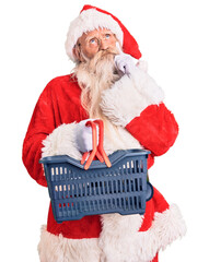 Old senior man with grey hair and long beard wearing santa claus costume holding shopping basket...