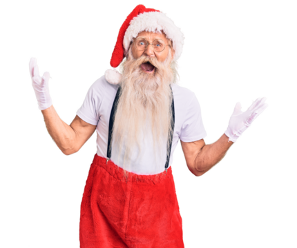 Old senior man with grey hair and long beard wearing santa claus costume with suspenders crazy and mad shouting and yelling with aggressive expression and arms raised. frustration concept.