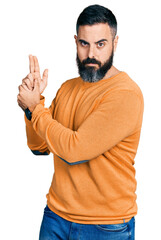 Fototapeta premium Hispanic man with beard wearing casual winter sweater holding symbolic gun with hand gesture, playing killing shooting weapons, angry face