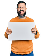 Hispanic man with beard holding blank empty banner sticking tongue out happy with funny expression.