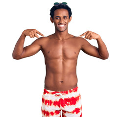Fototapeta premium African handsome man wearing swimsuit and sunglasses looking confident with smile on face, pointing oneself with fingers proud and happy.