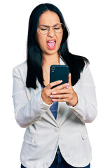 Fototapeta premium Beautiful hispanic woman with nose piercing using smartphone typing message angry and mad screaming frustrated and furious, shouting with anger. rage and aggressive concept.