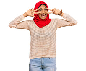 Naklejka premium Young african american woman wearing traditional islamic hijab scarf doing peace symbol with fingers over face, smiling cheerful showing victory