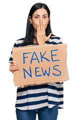 Beautiful young woman holding fake news banner covering mouth with hand, shocked and afraid for...