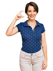 Young brunette woman with short hair wearing casual clothes smiling and confident gesturing with hand doing small size sign with fingers looking and the camera. measure concept.