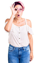 Fototapeta premium Young beautiful woman with pink hair wearing casual clothes and glasses doing ok gesture shocked with surprised face, eye looking through fingers. unbelieving expression.