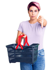 Young beautiful woman with pink hair holding supermarket shopping basket with angry face, negative sign showing dislike with thumbs down, rejection concept