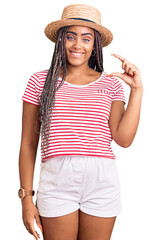 Young african american woman with braids wearing summer hat smiling and confident gesturing with hand doing small size sign with fingers looking and the camera. measure concept.