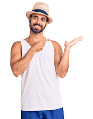 Young hispanic man wearing casual summer hat amazed and smiling to the camera while presenting with hand and pointing with finger.