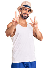 Young hispanic man wearing casual summer hat smiling looking to the camera showing fingers doing victory sign. number two.