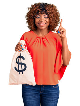 Young african american woman holding money bag with dollar symbol surprised with an idea or question pointing finger with happy face, number one