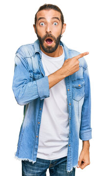 Attractive man with long hair and beard wearing casual denim jacket surprised pointing with finger to the side, open mouth amazed expression.