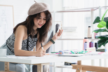 Asian attractive 30s woman fashion designer or dressmaker drawing sketch of new collection clothes on paper and smiling while working with tablet and tailoring equipments on desk at workplace studio
