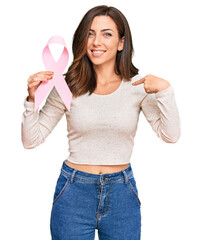 Young brunette woman holding pink cancer ribbon pointing finger to one self smiling happy and proud