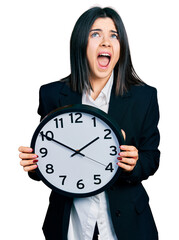 Young brunette woman with blue eyes holding big clock angry and mad screaming frustrated and furious, shouting with anger looking up.