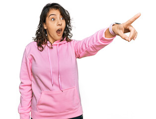 Young hispanic woman wearing casual sweatshirt pointing with finger surprised ahead, open mouth amazed expression, something on the front